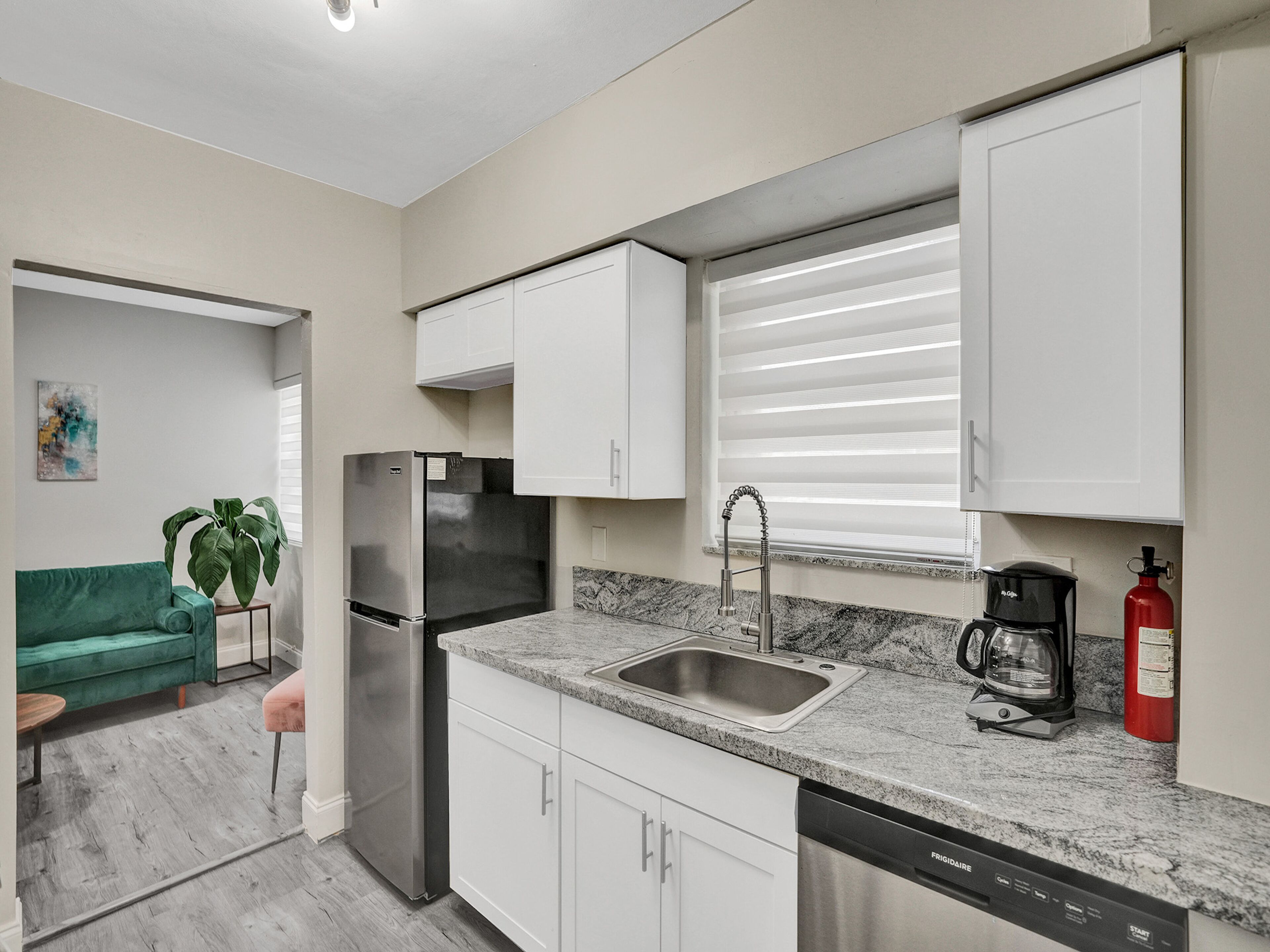 Cocina compacta con armarios blancos, refrigerador de acero inoxidable, un lavamanos, cafetera y extintor.