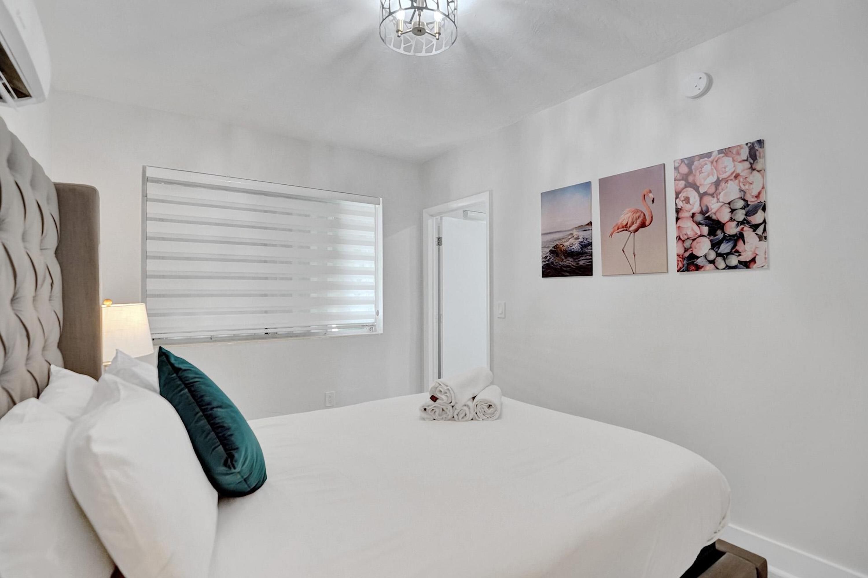 Une chambre à coucher avec un lit, une fenêtre avec des stores, une porte et trois tableaux accrochés au mur.