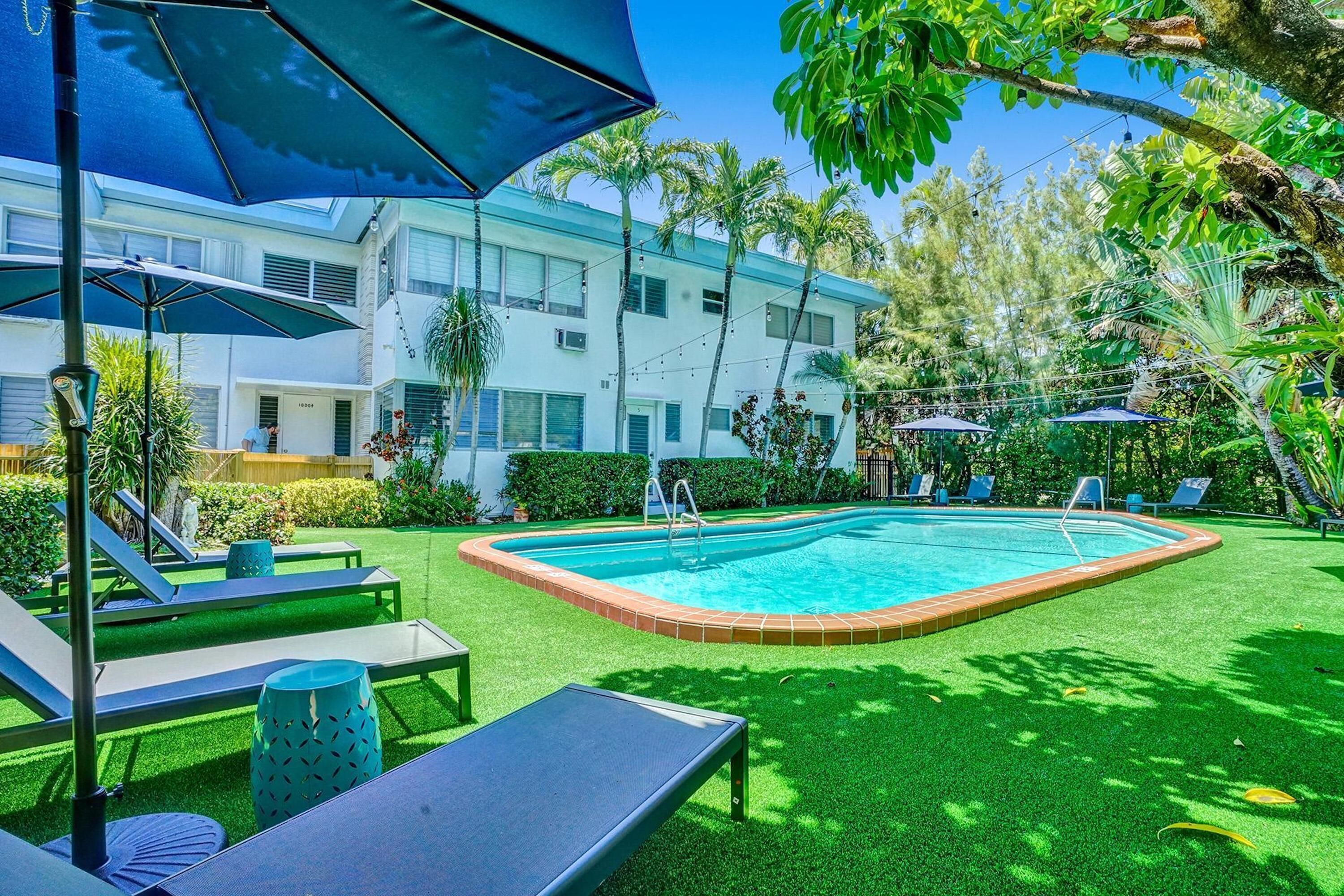 A pool area with lounge chairs and umbrellas, surrounded by greenery and a building in the background.