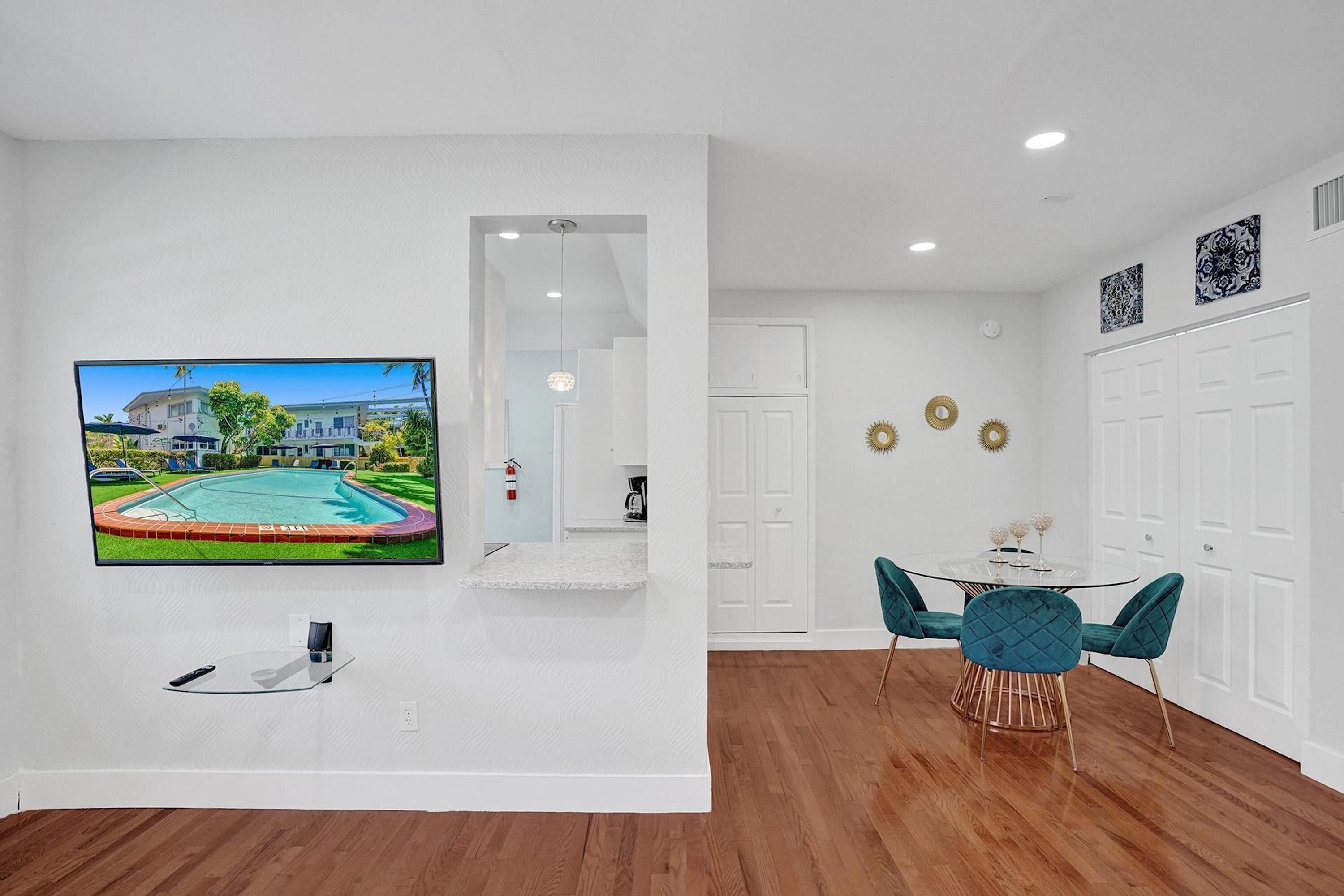 A modern living room with a flat-screen TV mounted on the wall, a dining area with a round table and teal chairs, and a wooden floor.