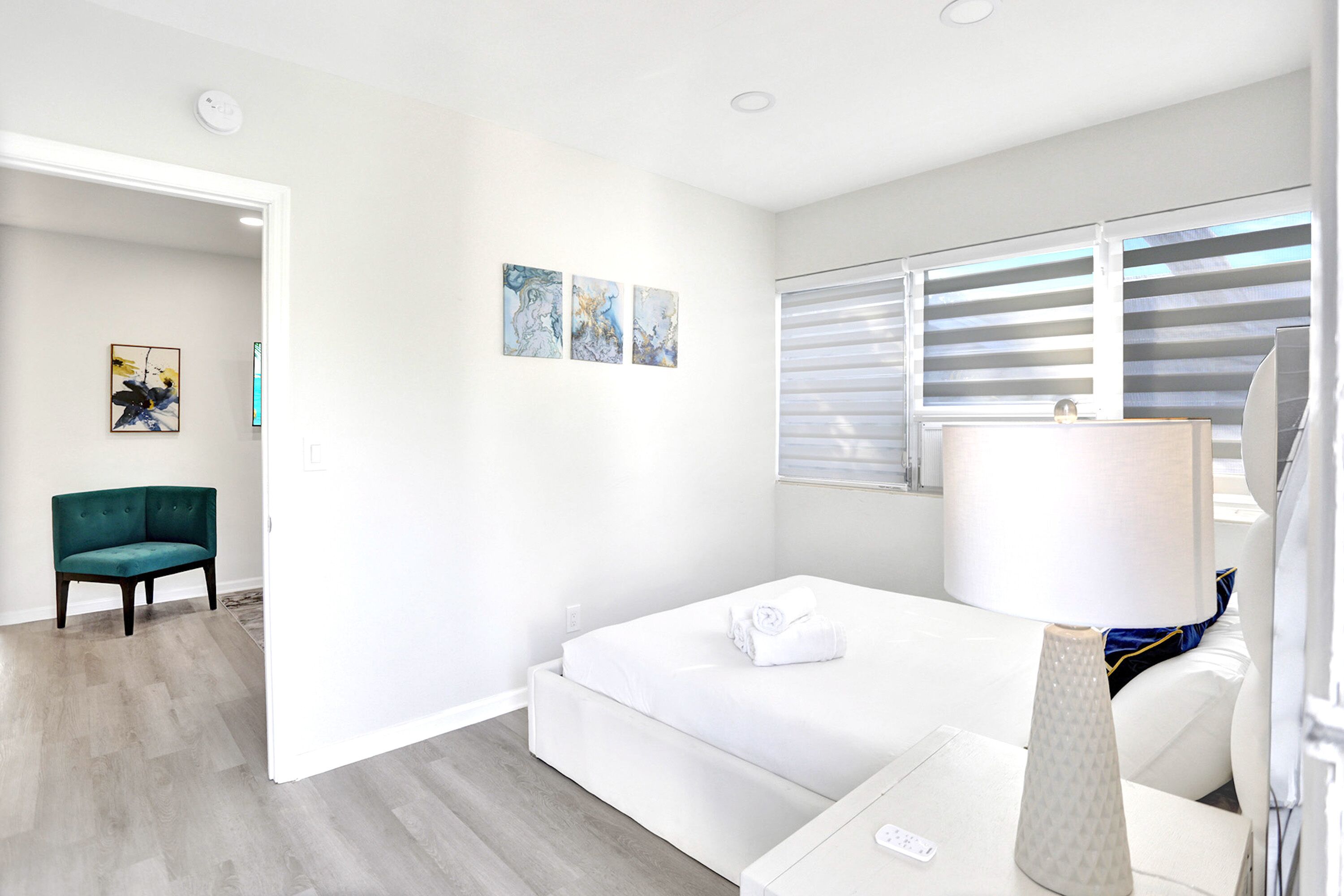 A modern hotel room with a bed, a bedside table with a lamp, a chair, and artwork on the wall.