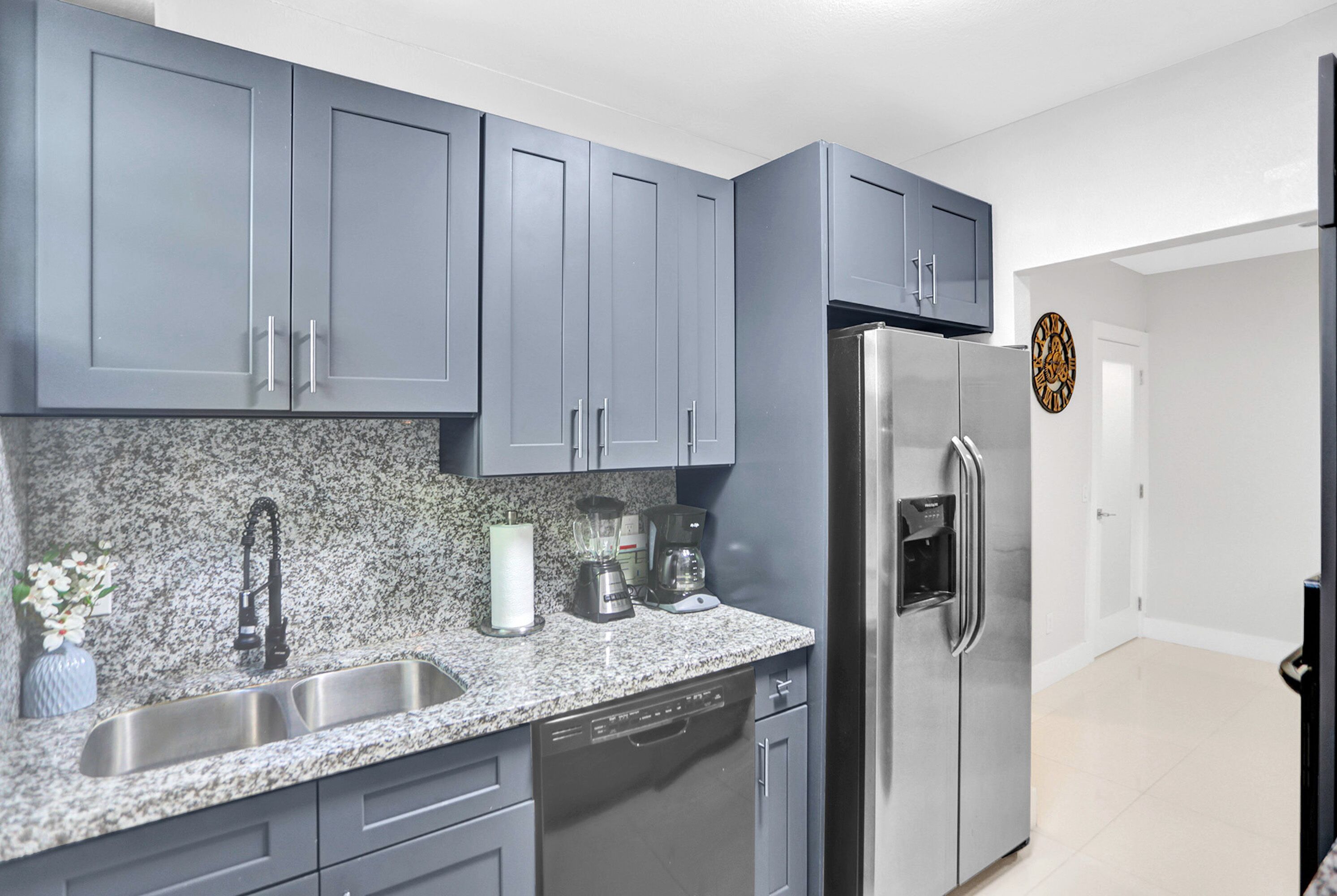 A modern kitchen with grey cabinets, granite countertops, a stainless steel refrigerator, and a dishwasher.