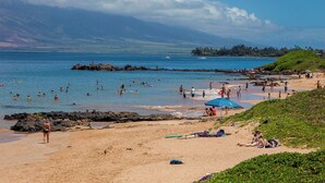 A beach with people swimming and sunbathing, a rocky outcrop, and a distant mountain range.