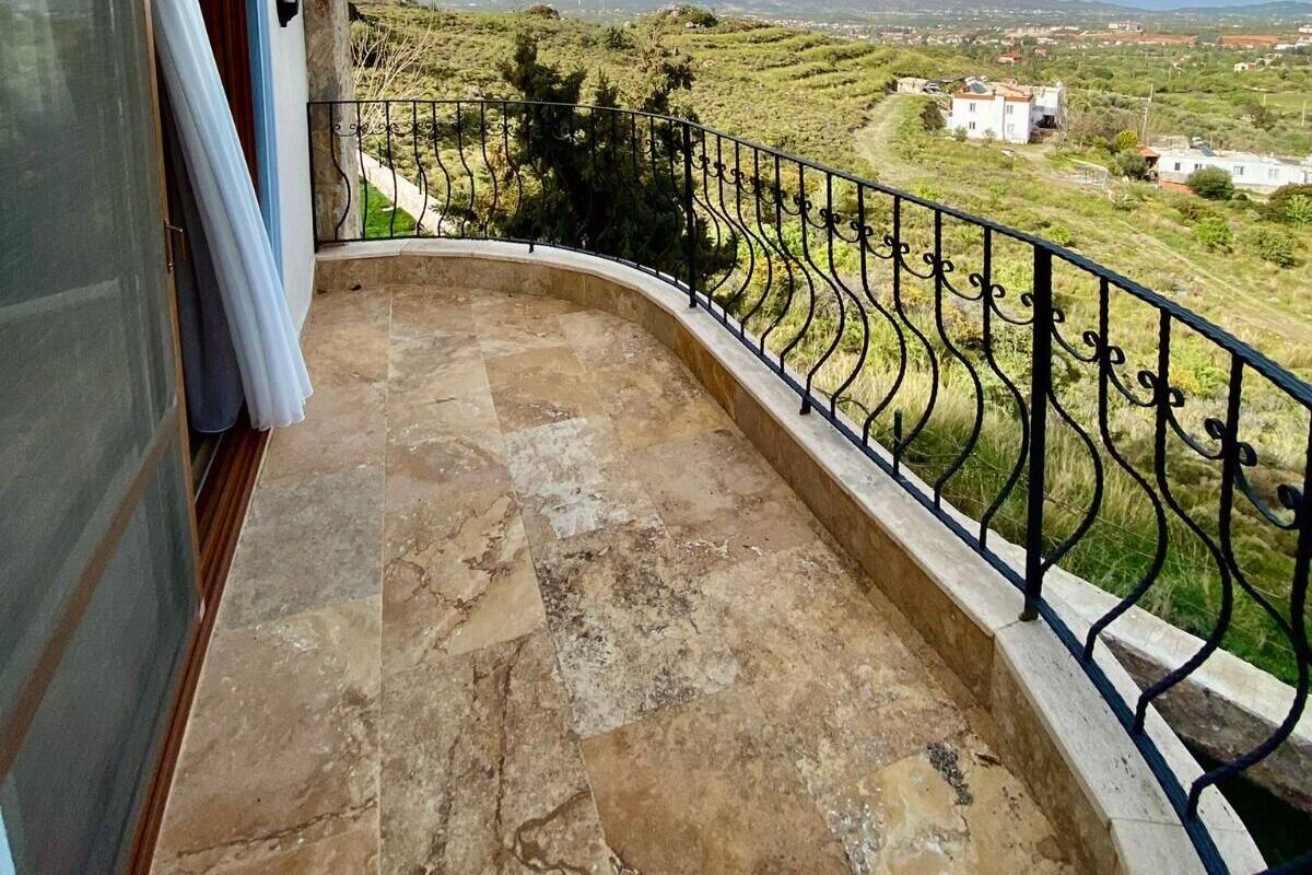 A balcony with a stone floor and metal railing, overlooking a green landscape with distant buildings.