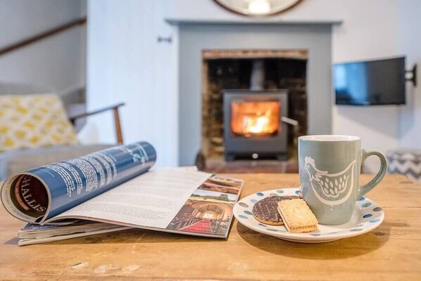 A cozy room with a fireplace, a table with a magazine, a mug, and biscuits. - 2 Riverside Cottages, Yoxford (Saxmundham)