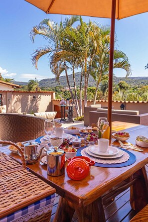 A breakfast setup with a table, wicker chairs, and a view of palm trees and hills. - Jardim Do Edém Piri (Pirenópolis)