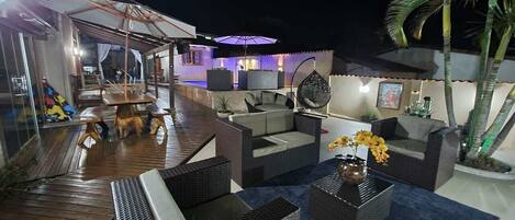 A well-lit outdoor patio area with wicker furniture, a dining table, and a bar area with a pool in the background.
