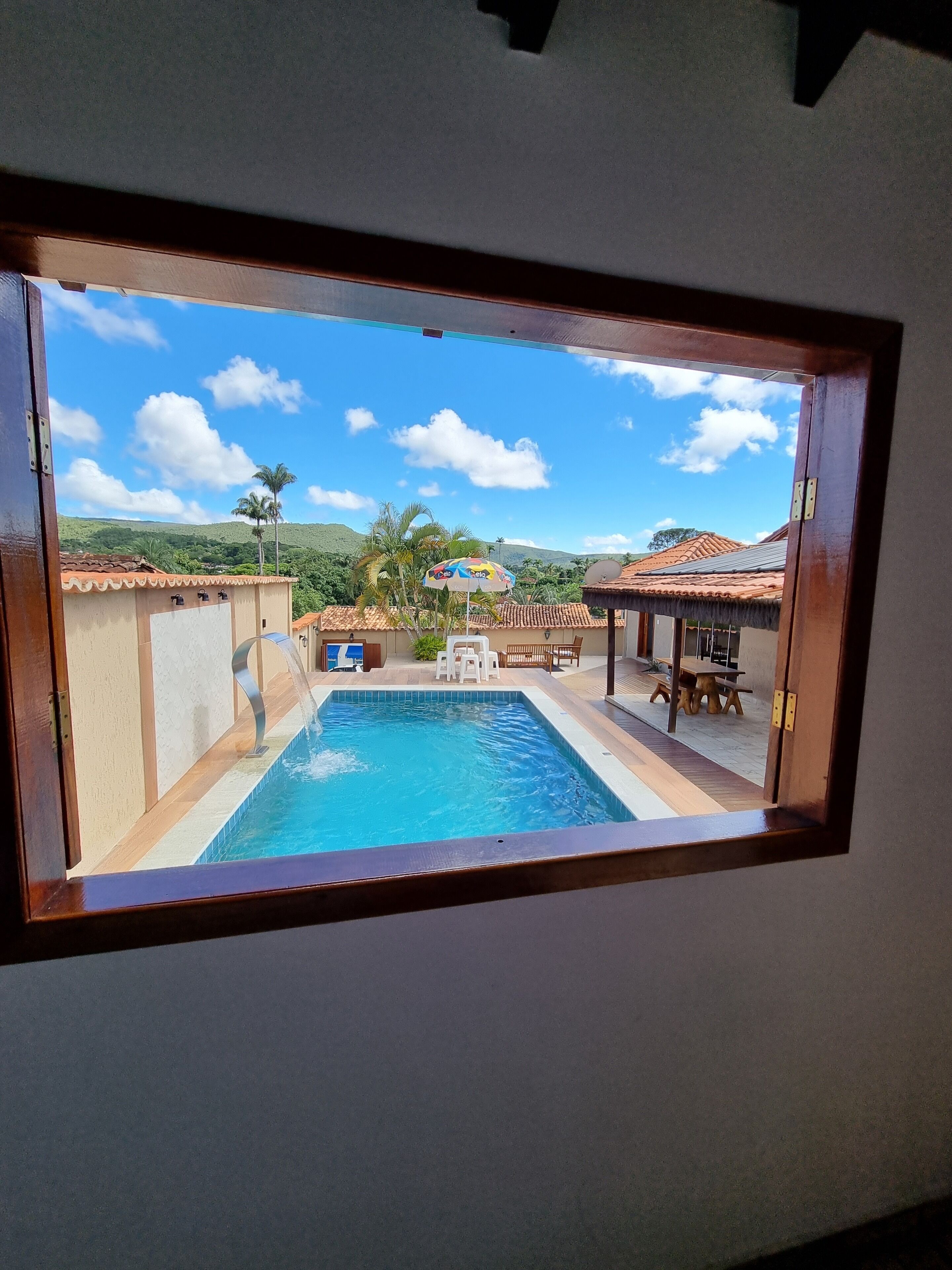 Une vue à travers une fenêtre sur un espace piscine avec une terrasse, du mobilier d’extérieur et un paysage montagneux en arrière-plan.