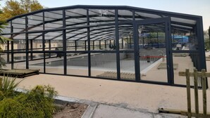 Un espacio al aire libre cubierto con estructura metálica y paneles de vidrio, rodeado de arena y vegetación.