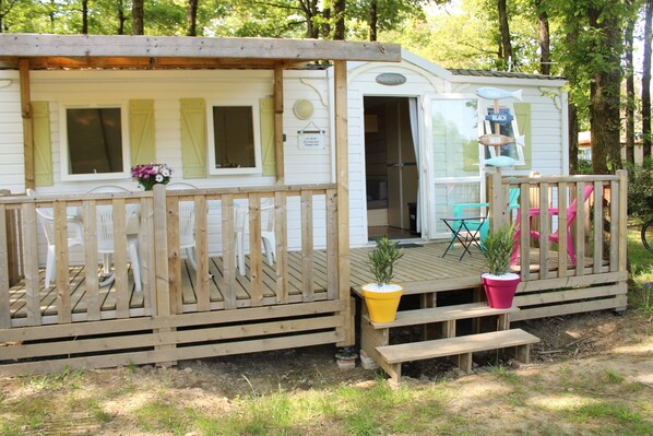 Une maison mobile dotée d’une terrasse, d’un petit espace extérieur aménagé pour s’asseoir, et d’un panneau indiquant les équipements disponibles, comme une plage et un bar.
