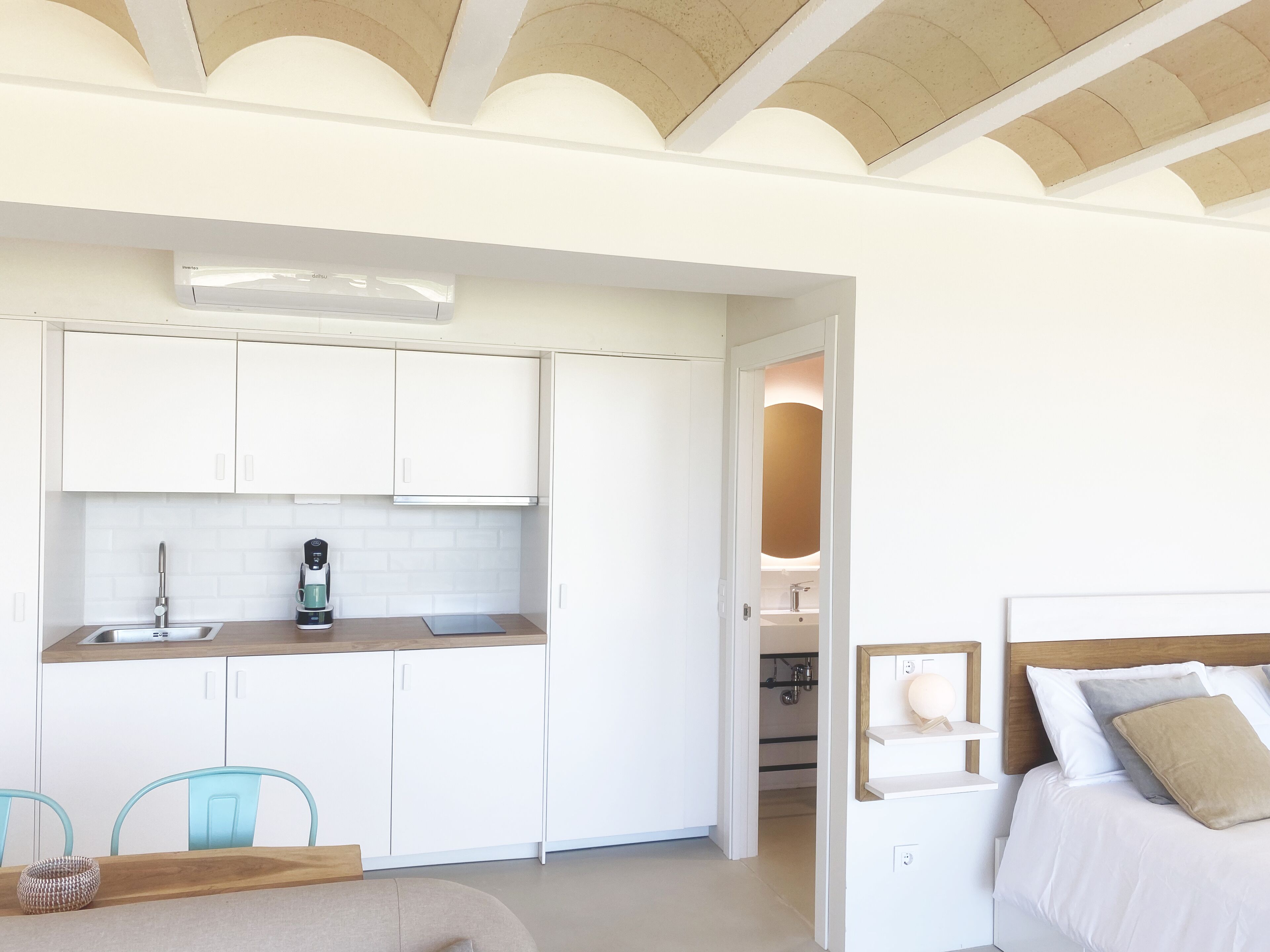Een moderne keuken met witte kasten, een koffieapparaat en een spoelbak. Er is een eethoek met een houten tafel en blauwe stoelen. Een slaapkamer met een bed en nachtkastje is zichtbaar door een open deur.