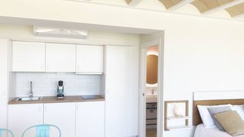 Een moderne keuken met witte kasten, een koffieapparaat en een spoelbak. Er is een eethoek met een houten tafel en blauwe stoelen. Een slaapkamer met een bed en nachtkastje is zichtbaar door een open deur.