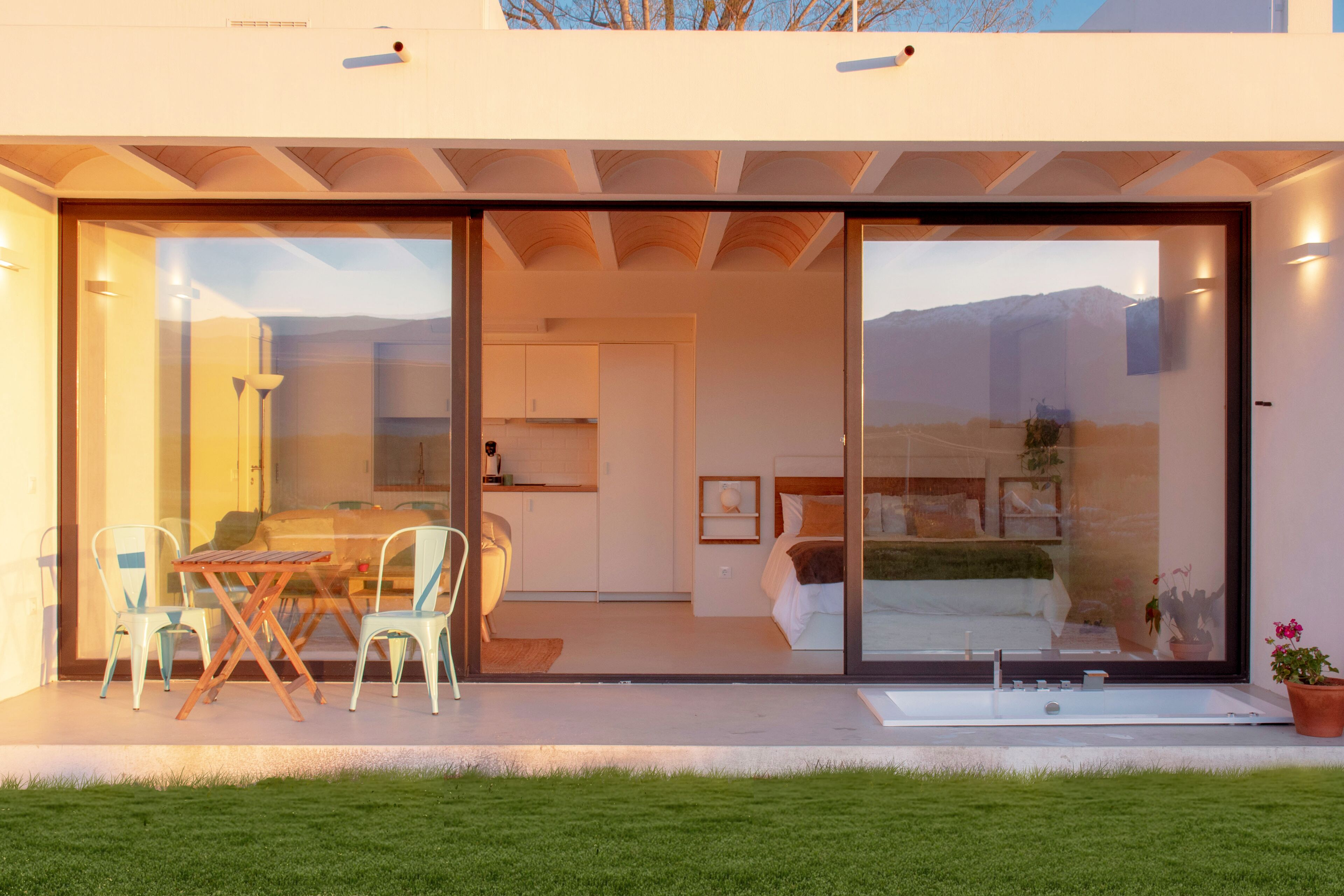 Une maison moderne dotée d’une porte vitrée qui donne sur un espace repas avec une table et des chaises, une cuisine et une chambre avec une grande fenêtre.