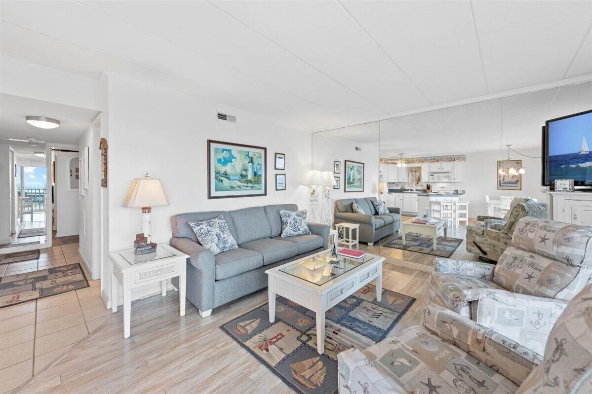 A spacious living room with a grey sofa, a coffee table, and a television. There are framed pictures on the walls and a kitchen area in the background.