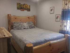A wooden bed with a patterned bedspread, a headboard with a painting, and a wooden nightstand. - Delaware River Retreat (Downsville)