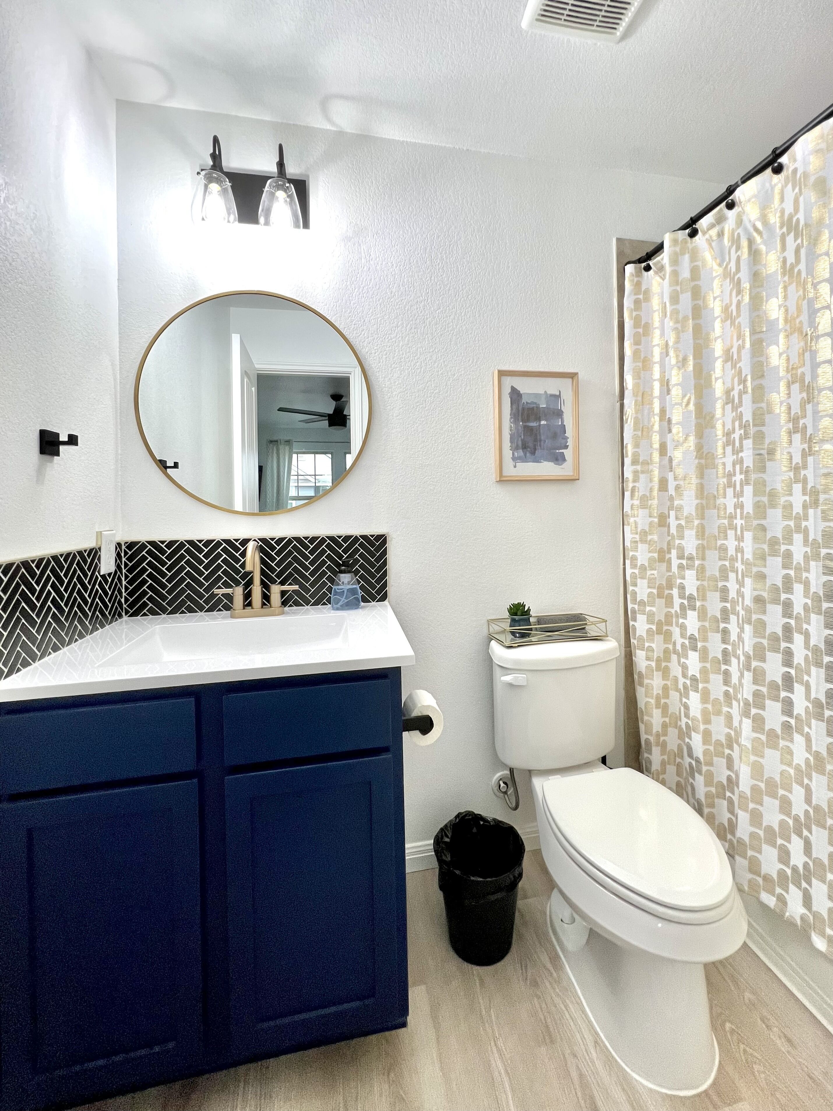 A bathroom with a white toilet, a blue vanity, a round mirror, and a patterned shower curtain.