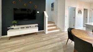 A modern living room with a flat-screen TV, a white entertainment unit, a staircase, and a dining area with a wooden table and chairs.