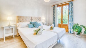Hotellihuone, jossa on kaksi sänkyä, puinen pääty, yöpöytä lampun kanssa, ikkuna verhoilla ja ruukkukasvi.