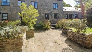 A stone building with multiple windows and a small garden area with various plants. - Barn Cottage (Penzance)