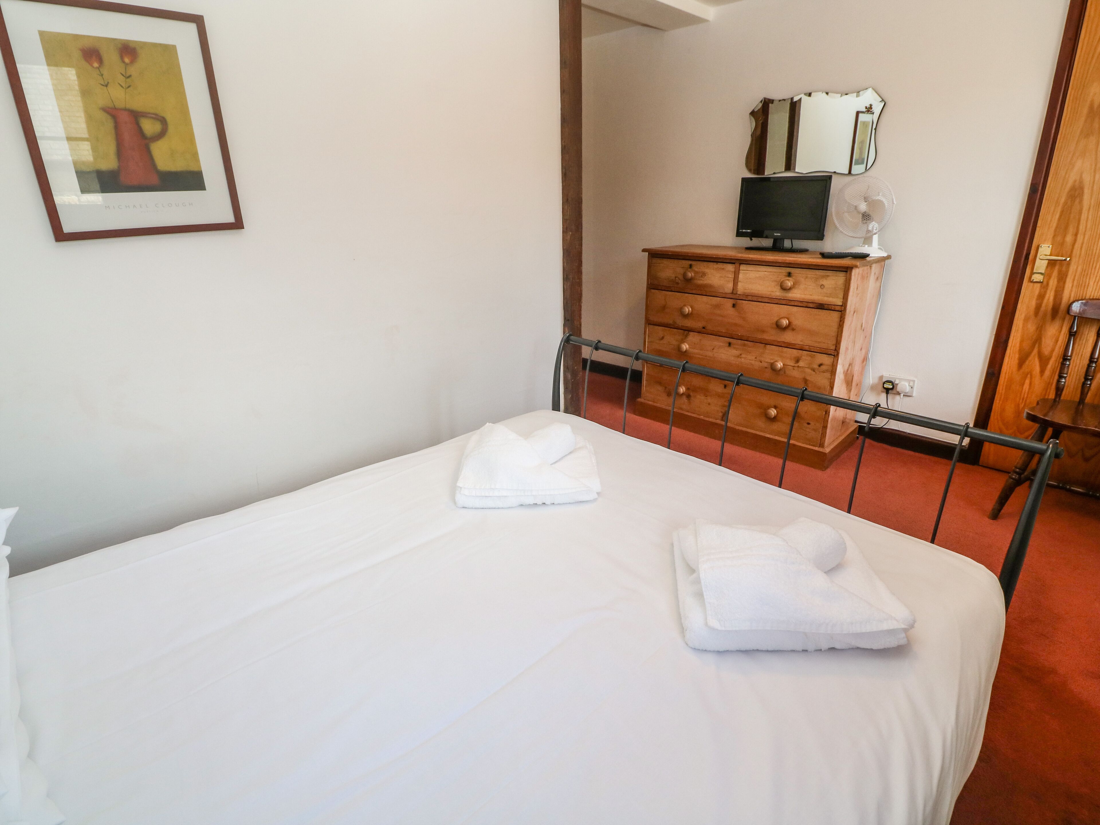 A neatly made bed with white linens and two folded towels, a wooden dresser with a television and fan, and a framed artwork on the wall.