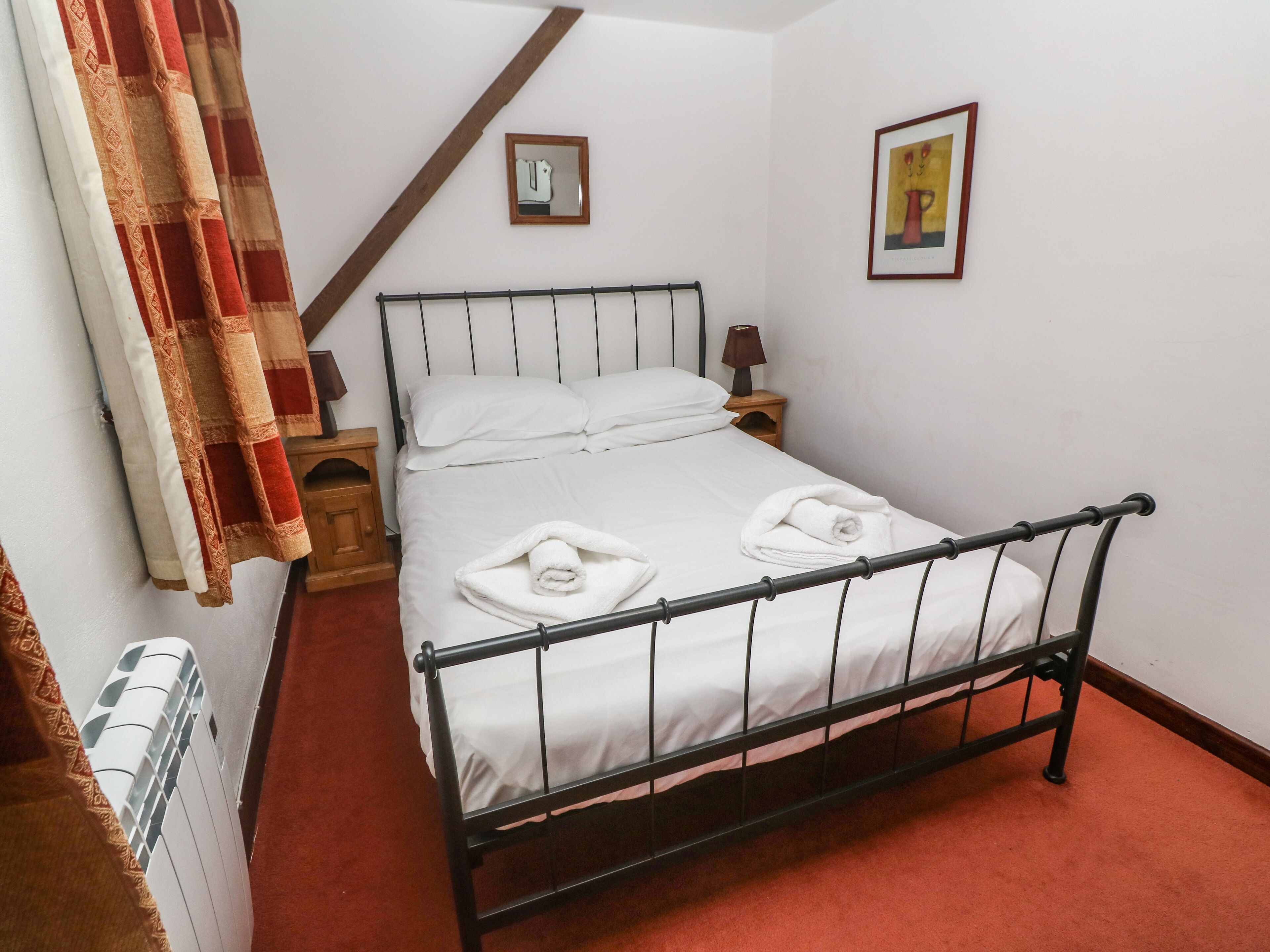 A single bed with white linens and two towels, a wooden nightstand with a lamp, a small framed mirror, and a painting on the wall.