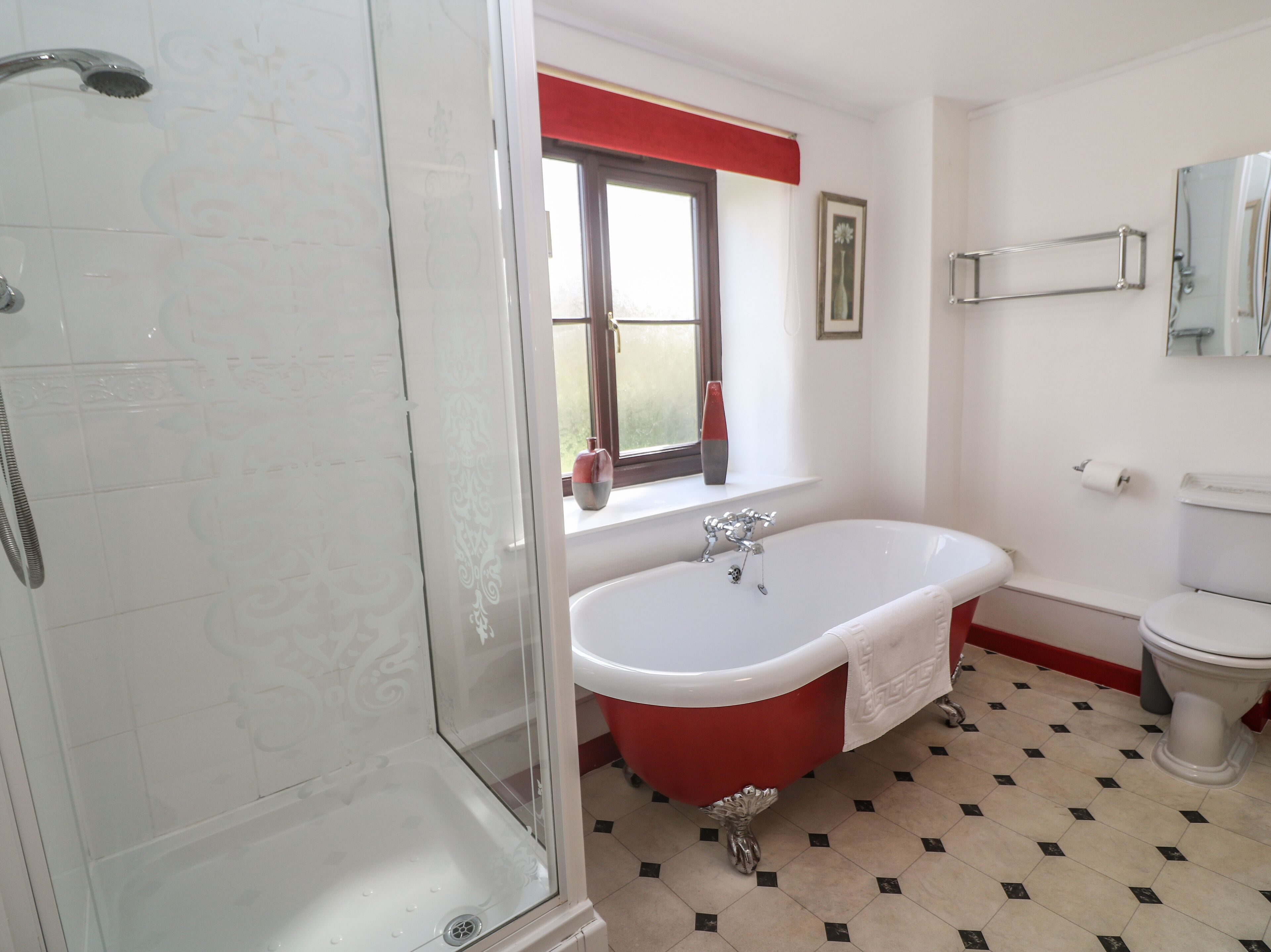 A bathroom with a glass shower enclosure, a red and white bathtub, a white toilet, and a window with a red valance.