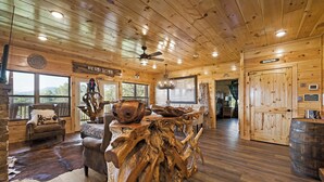 L’intérieur d’un chalet rustique, avec un plafond en bois, une table à manger faite de troncs d’arbres, des fauteuils en cuir et une grande fenêtre donnant sur le paysage extérieur.