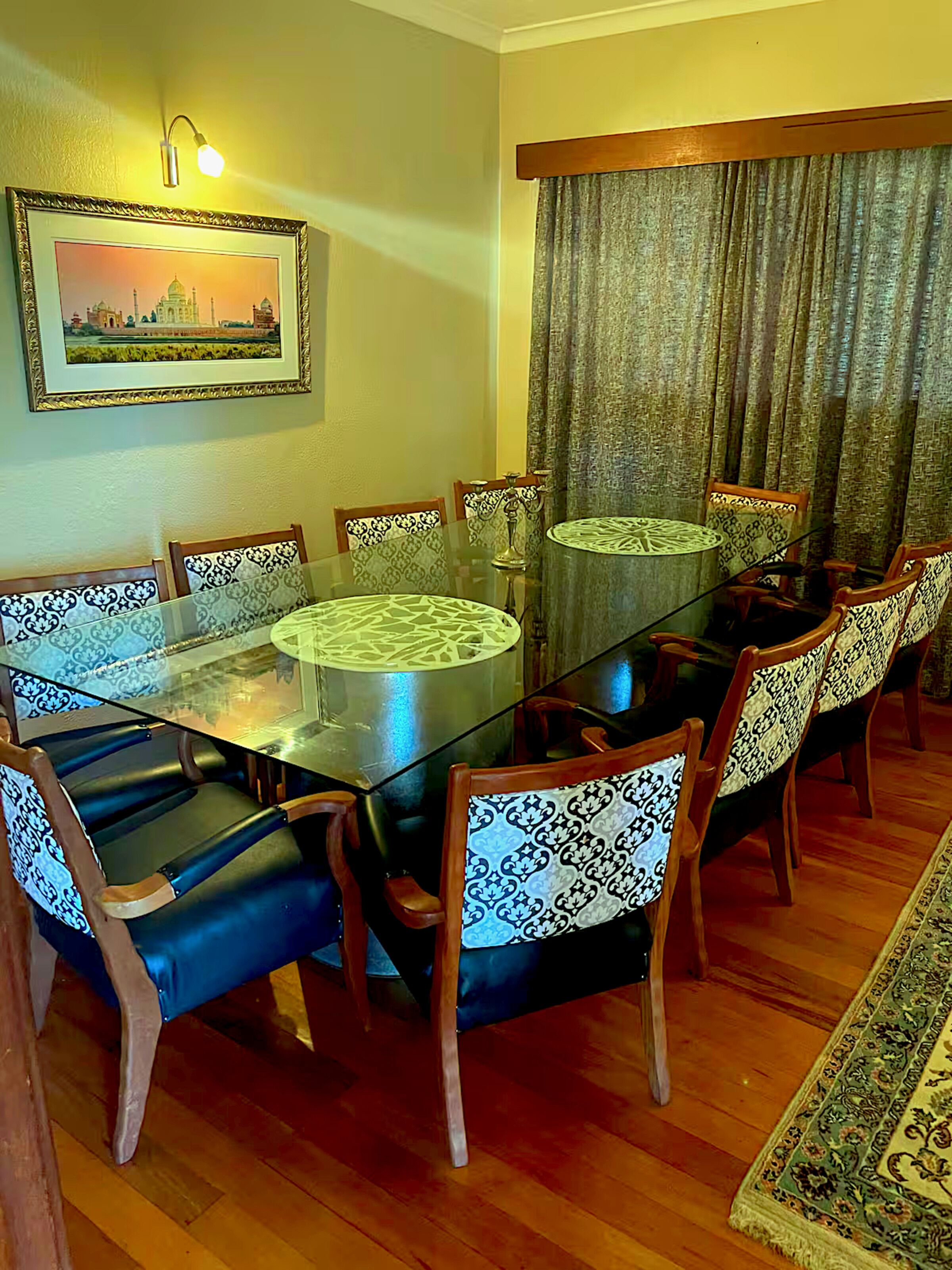 Une salle à manger avec une table en verre entourée de chaises à motifs, un tableau encadré au mur et un rideau à la fenêtre.
