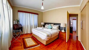 Une chambre avec un plancher en bois, un lit avec une literie blanche, une table de chevet avec une lampe, un bureau avec une chaise et une fenêtre avec des rideaux.