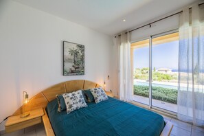 Une chambre à coucher avec un grand lit, une table de chevet, une lampe fixée au mur, un tableau et une vue sur un jardin, visible à travers une porte-fenêtre coulissante.