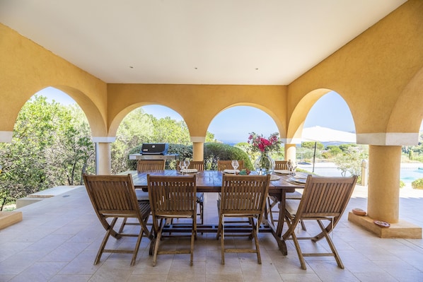 Un espace extérieur aménagé pour les repas, avec une table dressée pour quatre personnes, entourée de chaises en bois, et situé sous une structure couverte agrémentée d’arches.