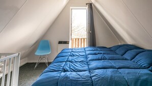 Ein Schlafzimmer mit schrägen Wänden, einem Bett mit blauer Bettwäsche, einem blauen Sessel und einem Fenster mit Vorhängen.