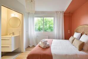 Quarto com cama, janela com cortinas, espelho redondo e banheiro branco com porta-toalhas.