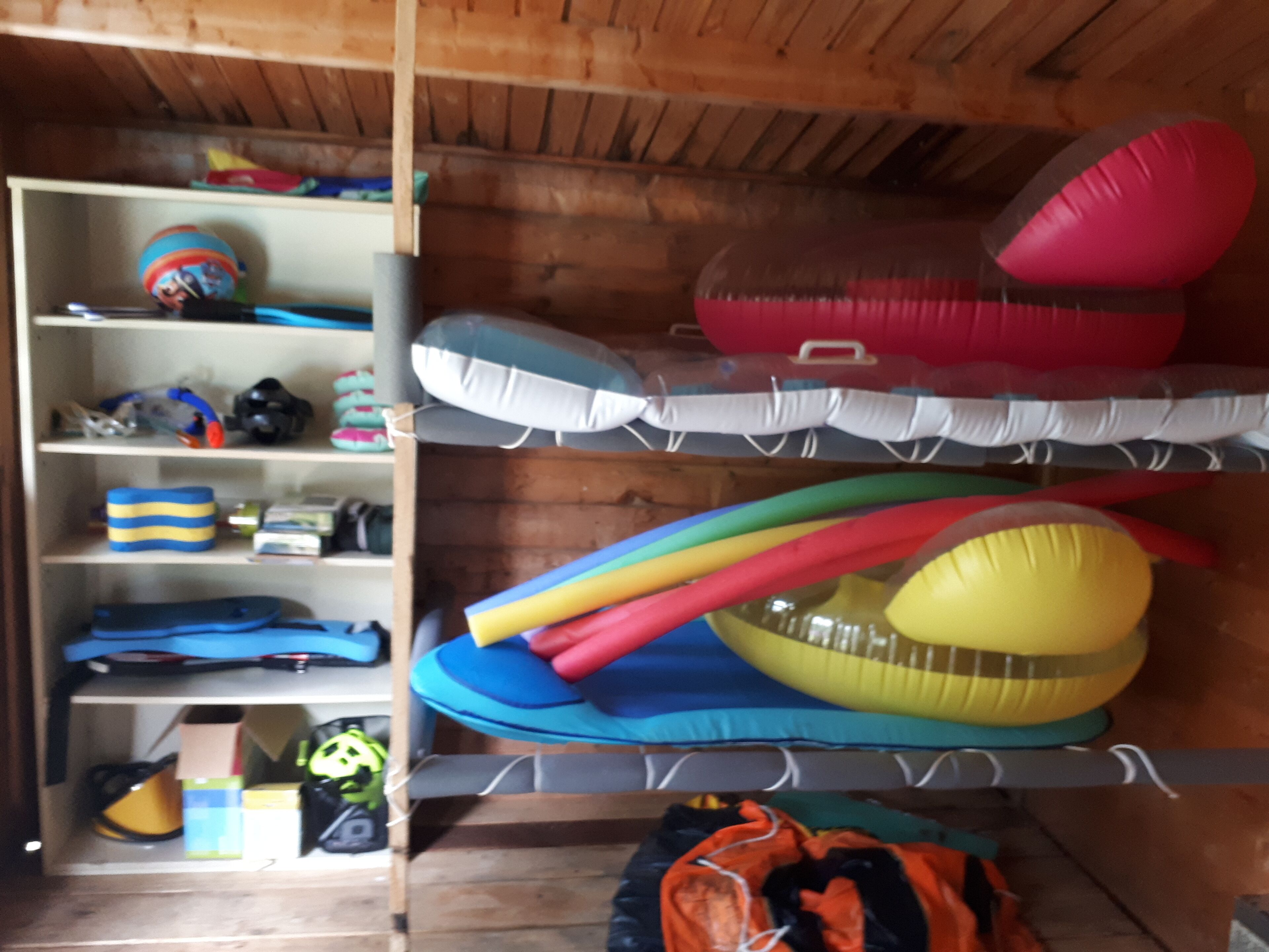 Ein Zimmer mit Holzwänden und einem Etagenbett mit bunten Matratzen. Es gibt Regale mit verschiedenen Gegenständen, darunter ein Helm und eine Tasche.