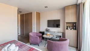 Een moderne hotelkamer met een bed, twee paarse fauteuils, een klein rond tafeltje met koffie en een tv die aan de muur is bevestigd, en een houten garderobe.