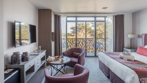 Een moderne hotelkamer met een groot bed, een bureau met een flatscreen televisie, twee stoelen, een kleine tafel met een vaas en een notitieboekje, en een balkon met uitzicht op bomen.