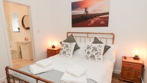 A neatly made bed with a metal frame, two bedside tables with lamps, a painting on the wall, and a bathroom visible through an open door. - The Bakery (Belford)
