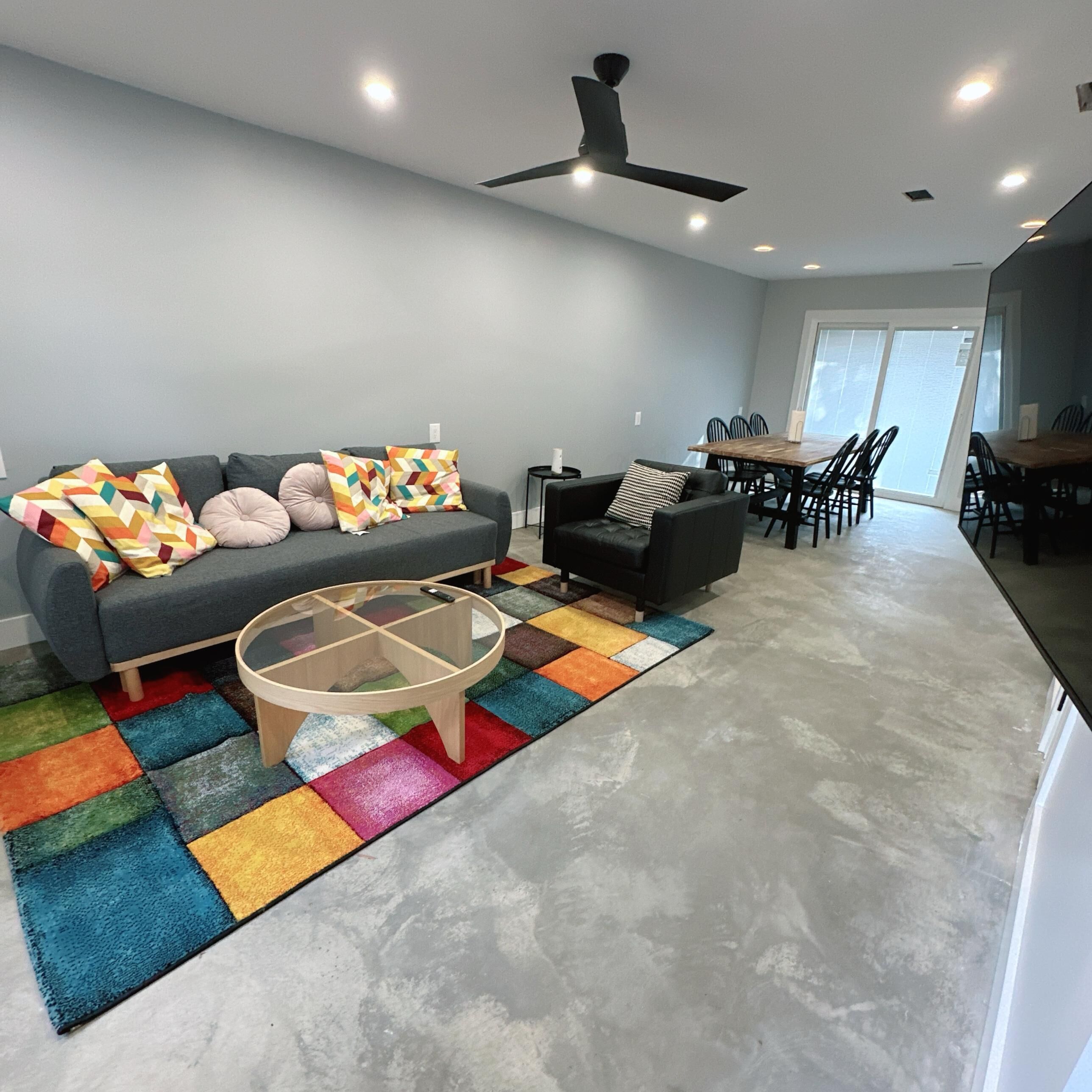 A modern living room with a grey sofa, a black armchair, and a glass coffee table. There is a colorful rug on the floor and a ceiling fan above.