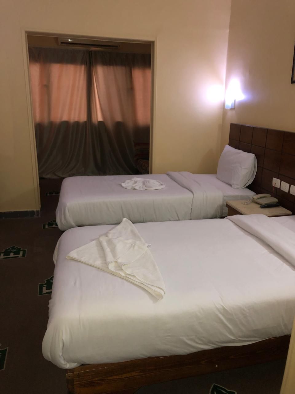 A hotel room with two single beds, a bedside table with a lamp, and a window with curtains.