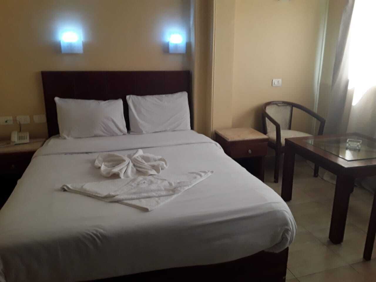 Una habitación de hotel con una cama bien hecha, una mesita de noche con una lámpara, una silla y una pequeña mesa con un vaso.