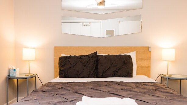Un letto rifatto con cura, una testiera marrone scuro e cuscini, due abat-jour e uno specchio sopra il letto.