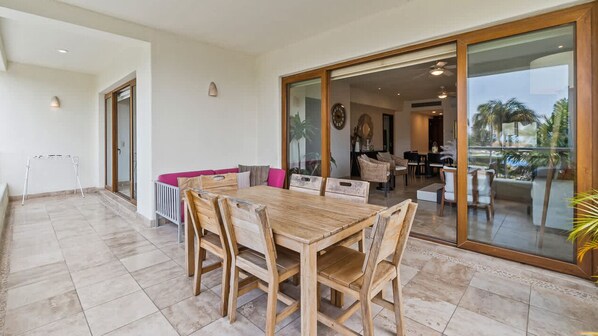 Un patio amplio con una mesa y sillas de madera, un banco acolchado y un ventanal corredero que da a un espacio interior con una mesa de comedor y sillas.