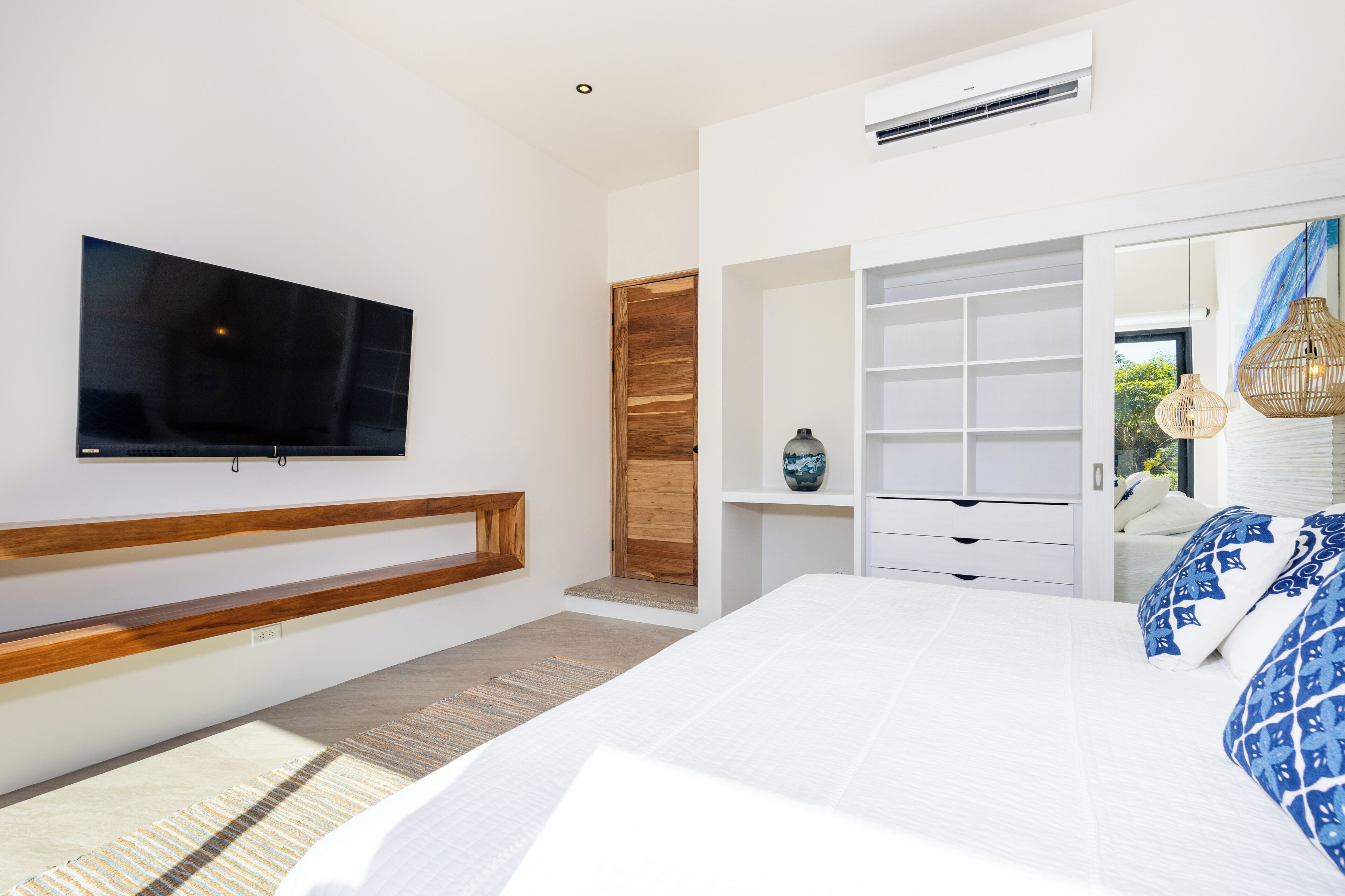 Un dormitorio con una cama grande, un televisor fijado a la pared, un banco de madera, un armario y un espejo que refleja la vista de una ventana.