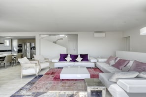 Un salon moderne avec un ensemble de canapés blancs, une table basse et un tapis à motifs. Un escalier mène à un autre niveau.