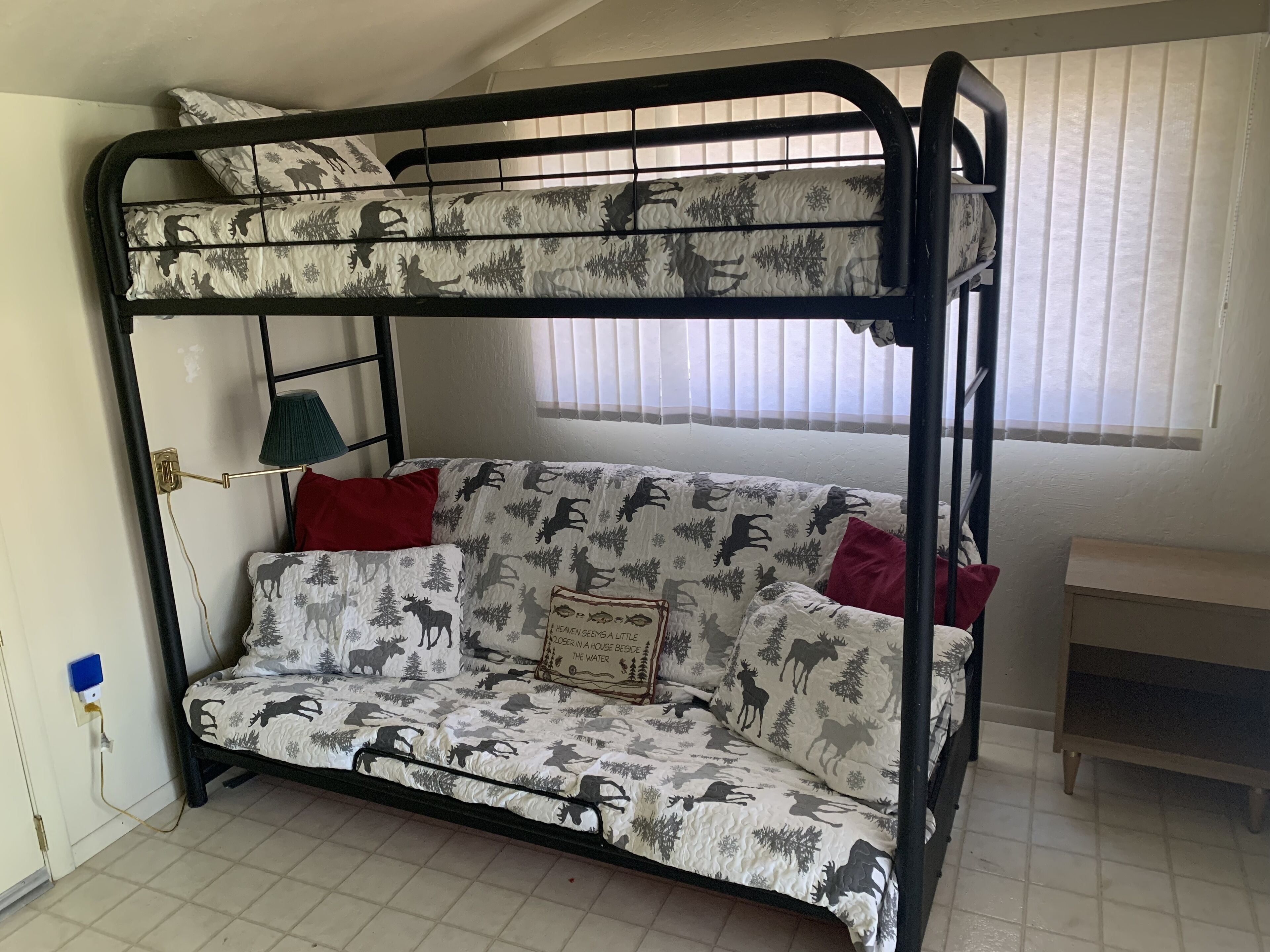 Un lit superposé dont le lit du bas est doté d’un matelas et de coussins à motifs, d’une lampe et d’une fenêtre avec des stores.