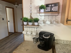 Een keuken met een koffiezetapparaat, mokken op een rek, potplanten en een bordje met de tekst 'GOOD FOOD GOOD MOOD'.