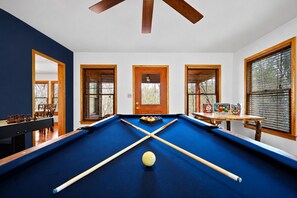 Une table de billard recouverte d’un feutre bleu, des queues de billard en bois et une bille de billard jaune. Il y a trois portes avec des cadres en bois et des panneaux de verre, et un ventilateur de plafond au-dessus.