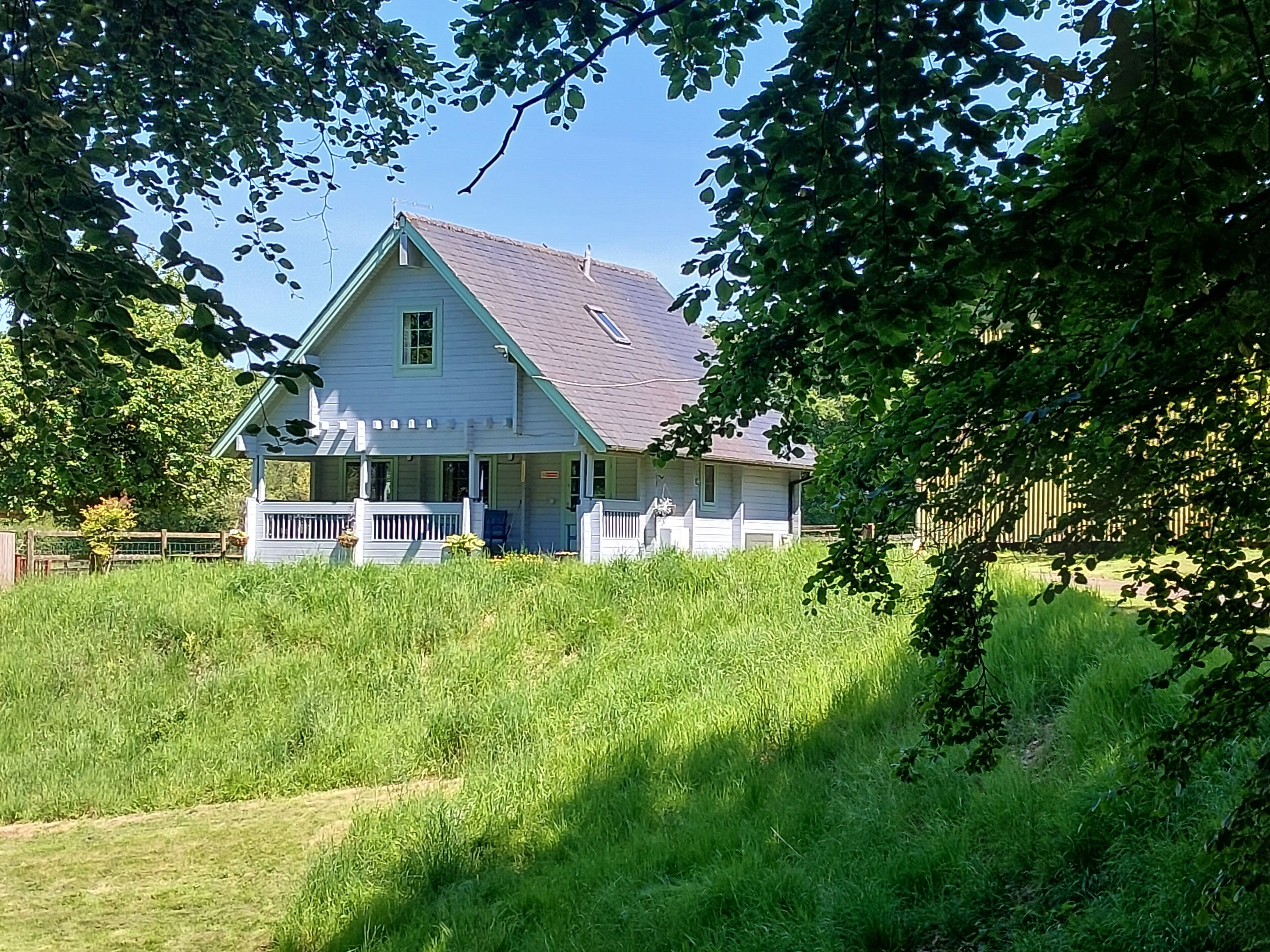 Une maison à deux étages avec un porche, entourée d’herbes hautes et d’arbres.