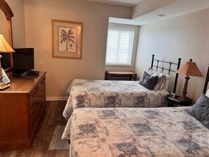 A bedroom with two beds, a desk with a television, a framed picture on the wall, and a window with blinds. - Sunrise Pointe 11F (North Myrtle Beach)