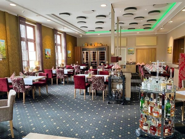 A restaurant with multiple tables and chairs, a bar area with bottles, and a large window with curtains. - Hotel Am Schloss Aurich Schlossresidenz (Aurich)